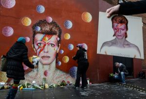 A man holds a copy of the David Bowie album "Aladdin Sane" as floral tributes are left beneath a mural of the British singer painted by Australian street artist James Cochran, aka Jimmy C, following the announcement of Bowie's death, in Brixton, south London, on Jan. 11, 2016. (Credit: AFP PHOTO / CHRIS RATCLIFFE) RESTRICTED TO EDITORIAL USE, MANDATORY MENTION OF THE ARTIST UPON PUBLICATION, TO ILLUSTRATE THE EVENT AS SPECIFIED IN THE CAPTION / AFP / CHRIS RATCLIFFE (Photo credit should read CHRIS RATCLIFFE/AFP/Getty Images)