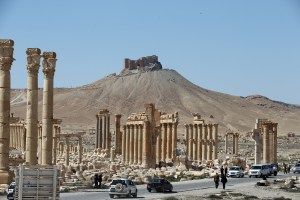 A picture shows people walking near the remains of Arch of Triumph, also called the Monumental Arch of Palmyra, on March 31, 2016, in the ancient Syrian city of Palmyra, which was destroyed by jihadists of the Islamic State group in 2015. (Credit: Joseph Eid/AFP/Getty Images)