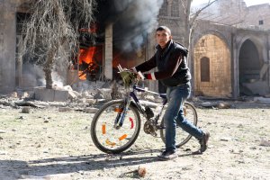 A Syrian youth pushes his bicycle past a burning house on Nov. 19, 2016, following a reported air strike on a rebel-held neighborhood of Aleppo. (Credit: Ameer Alhalbi/AFP/Getty Images)