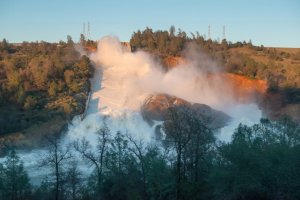 The heavy flows from the 3,000-foot main Oroville dam spillway continues to push debris into the turbid Feather River as the concrete span further eroded and the jagged fracture in its midsection sends water flowing over an adjacent hillside. (Credit: Florence Low / California Department of Water Resources)