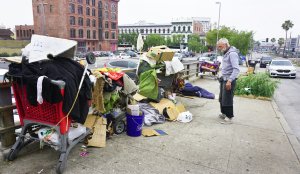 A homeless man stands before his belongings at his encampment on a downtown Los Angeles sidewalk as vehicles pass by on June 7, 2017. (Credit: Frederic J. Brown / AFP / Getty Images)