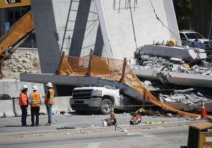 Workers stand near a crushed vehicle as law enforcement and members of the National Transportation Safety Board investigate the scene where a pedestrian bridge collapsed a few days after it was built, March 16, 2018, in Miami, Florida. (Credit: Joe Raedle / Getty Images)