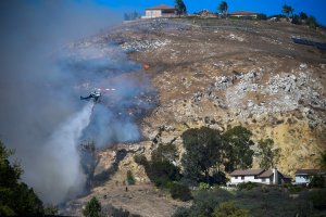 A helicopter drops water over burning embers on a hillside overlooking homes in West Hills, near Malibu on Nov. 11, 2018, as the battle to control the Woolsey Fire continued. (Credit: Frederic J. Brown/AFP/Getty Images)